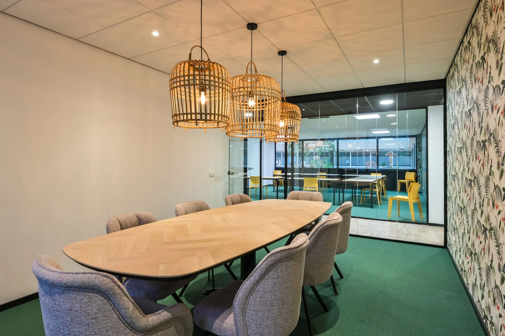 Vergaderruimte aan de Havenweg 24 met een ovale houten tafel, gestoffeerde stoelen en rotan hanglampen, grenzend aan een ruimte met gele stoelen achter een glazen wand.
