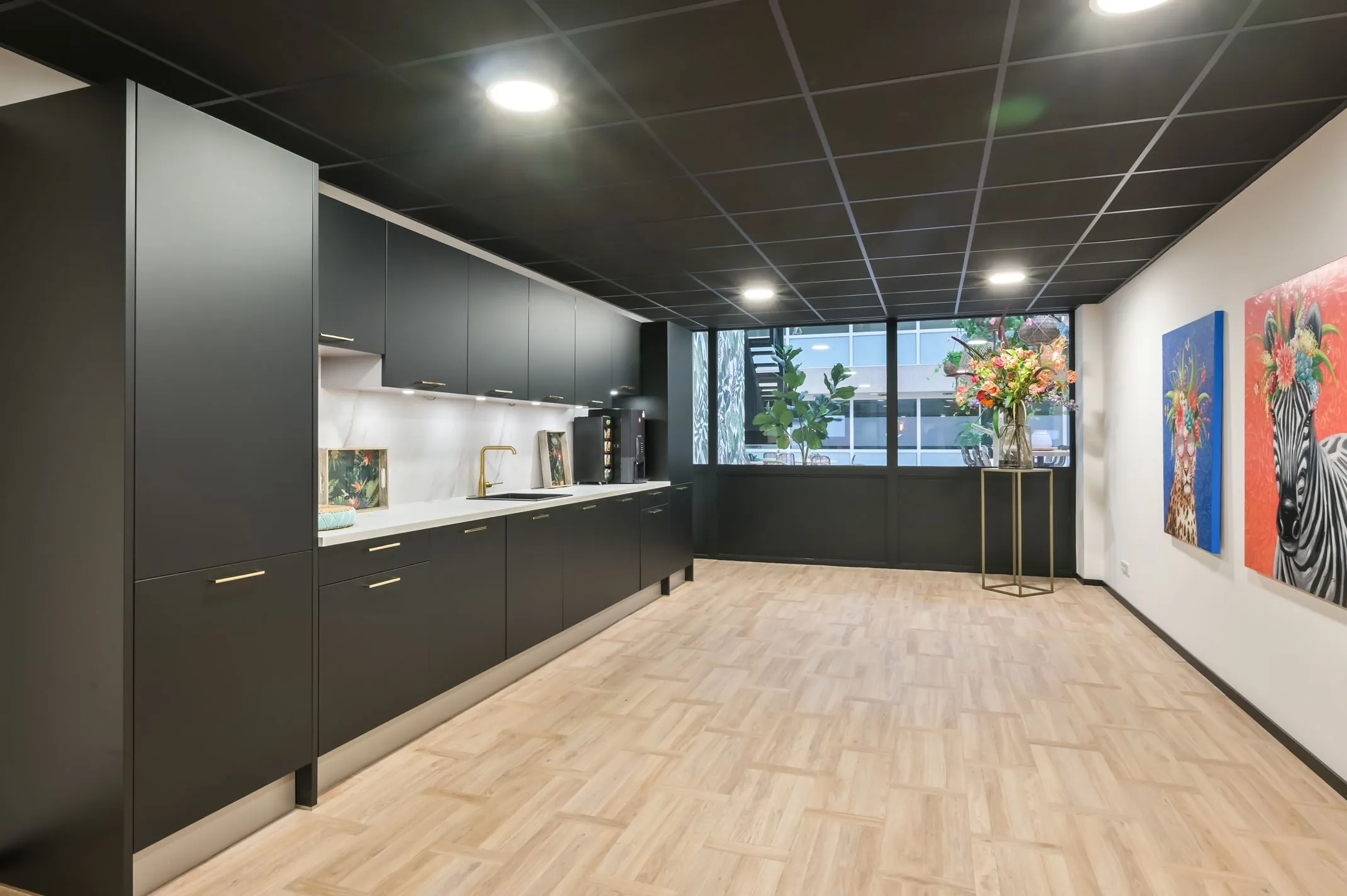 Moderne pantry met zwarte keukenkasten, houten vloer, kleurrijke dierenkunst aan de muur en grote vaas met bloemen bij Havenweg 24.