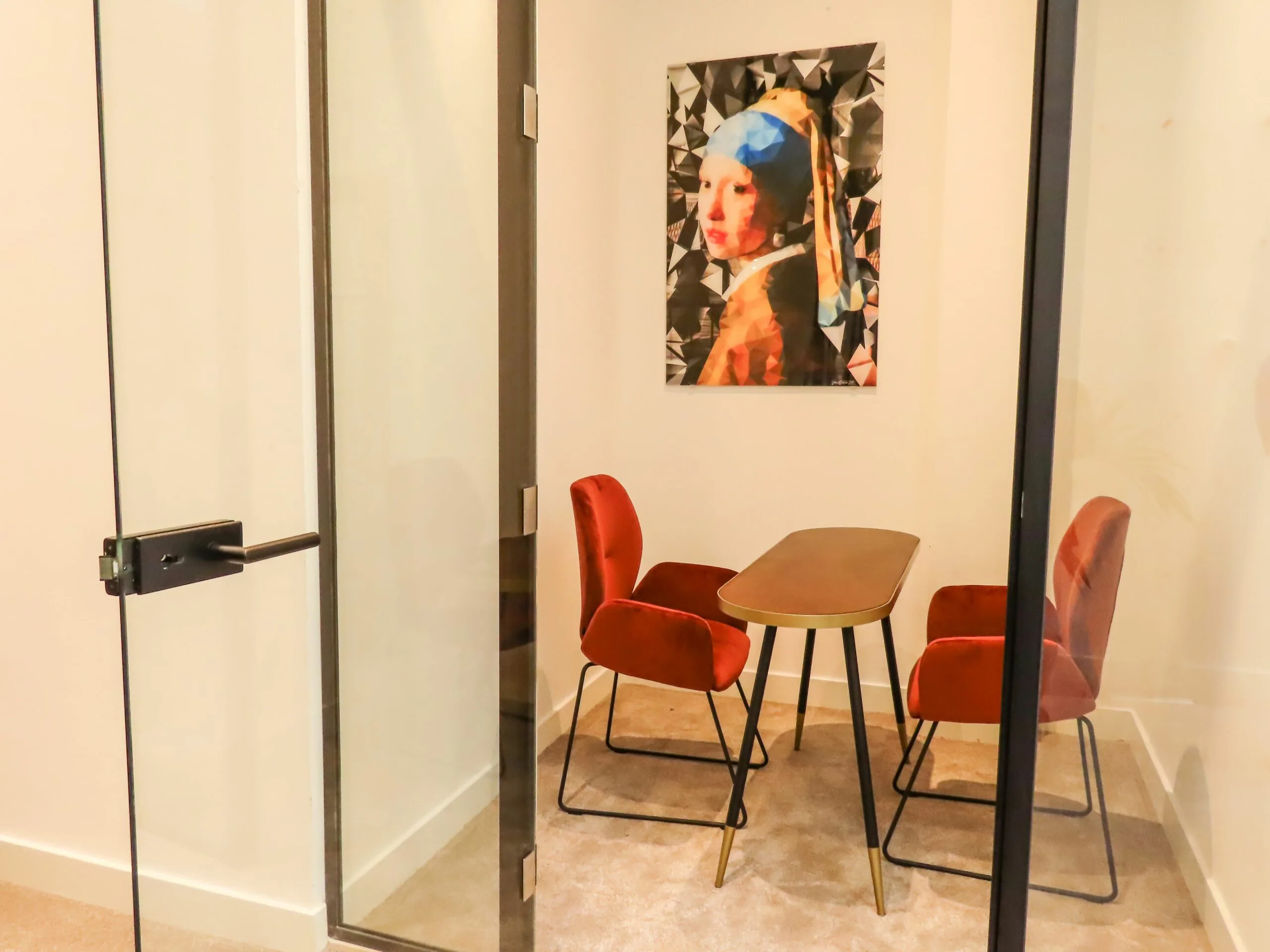 Kleine vergaderruimte met twee rode stoelen, een ovale tafel en een modern schilderij van het Meisje met de parel aan de muur.