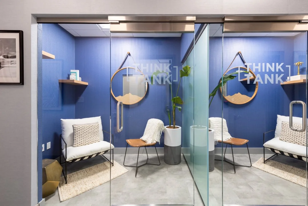 Moderne, glazen vergaderruimtes met blauwe muren en de tekst "Think Tank" op de deuren, ingericht met stoelen, planten en decoratieve elementen.