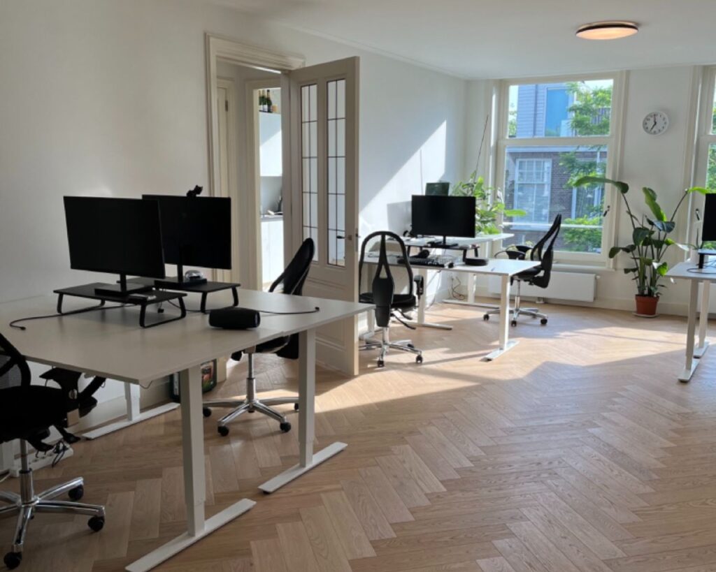 Licht en modern kantoor met meerdere werkplekken, bureaustoelen, computerschermen en grote ramen met planten.