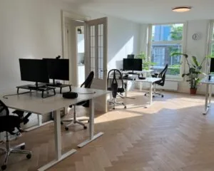 Licht en modern kantoor met meerdere werkplekken, bureaustoelen, computerschermen en grote ramen met planten.