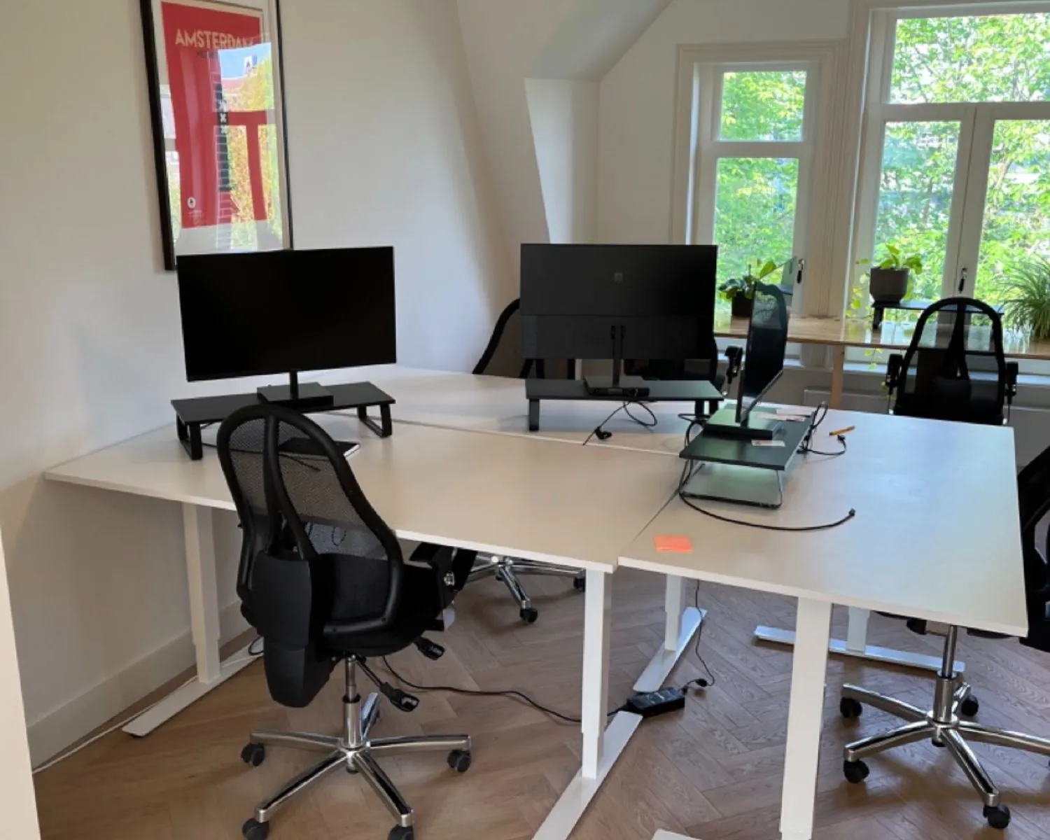 Modern kantoor met vier bureaus, computerschermen, ergonomische stoelen en een groot raam met uitzicht op groen.