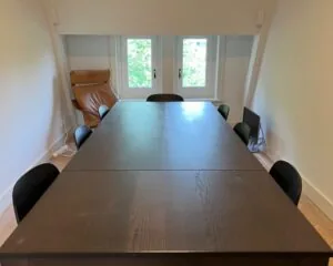 Vergaderruimte met een donkere houten tafel, acht zwarte stoelen, een bruine loungestoel en een computerscherm bij het raam.