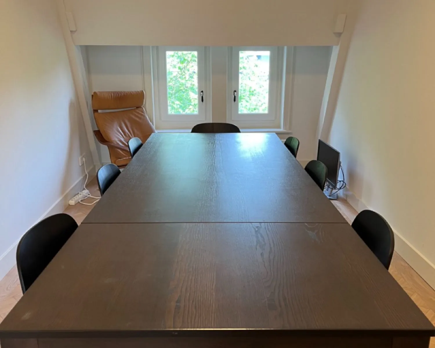 Vergaderruimte met een donkere houten tafel, acht zwarte stoelen, een bruine loungestoel en een computerscherm bij het raam.