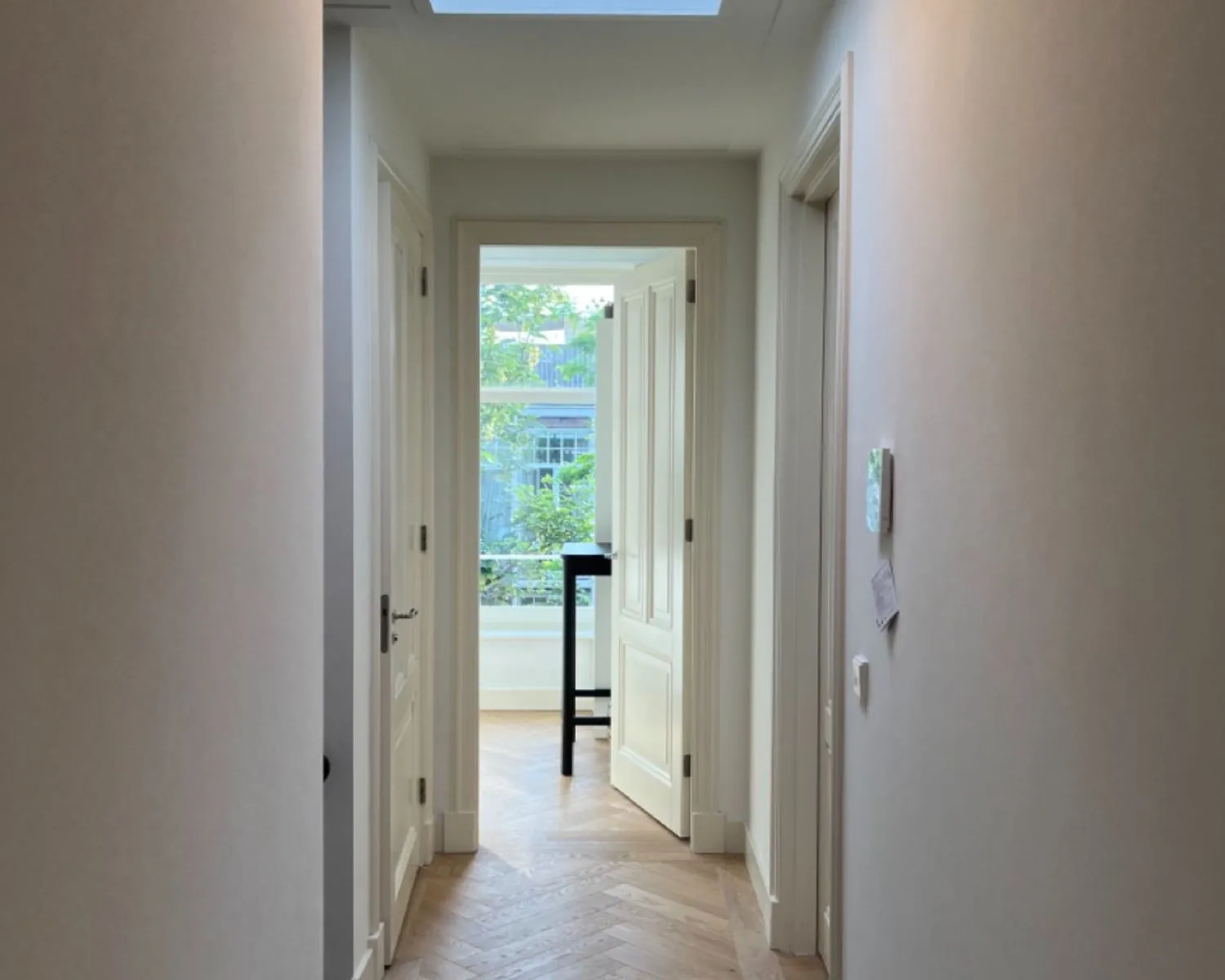 Lichte gang met houten visgraatvloer en uitzicht op een openstaande deur naar een kamer met een hoog raam en zwart tafeltje.
