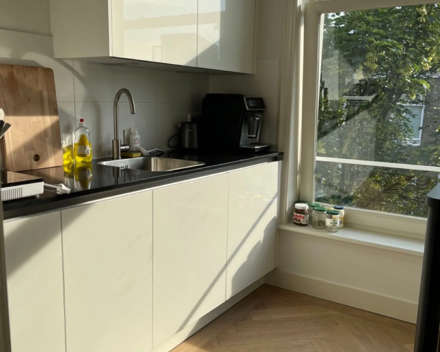 Moderne keuken met wit glanzend aanrecht, zonlicht door het raam en potjes op de vensterbank.