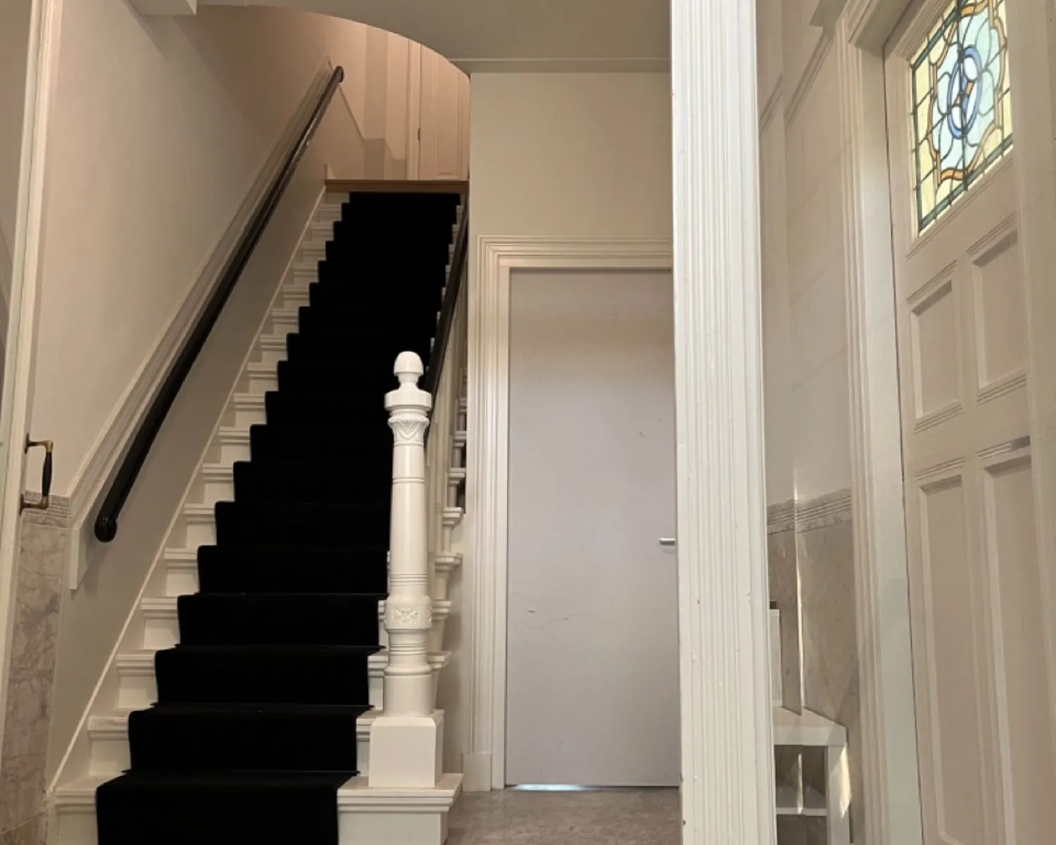 Trap met zwarte loper en wit houten hekwerk in een klassieke hal met paneeldeur en glas-in-loodraam.