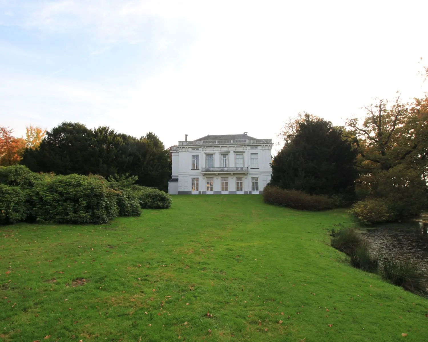 Wit landhuis met symmetrische gevel, omgeven door groen gras en bomen, gezien vanaf de tuinzijde.