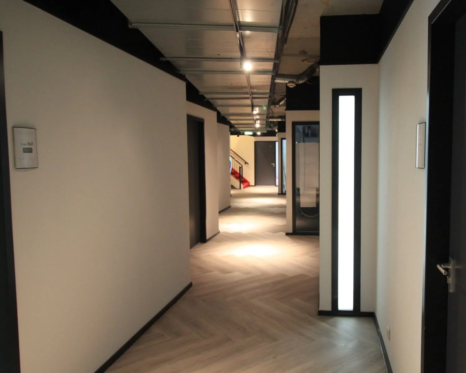 Verlichtte gang met lichte houten vloer, zwarte plafondaccenten en zicht op een rode glijbaan in de verte.