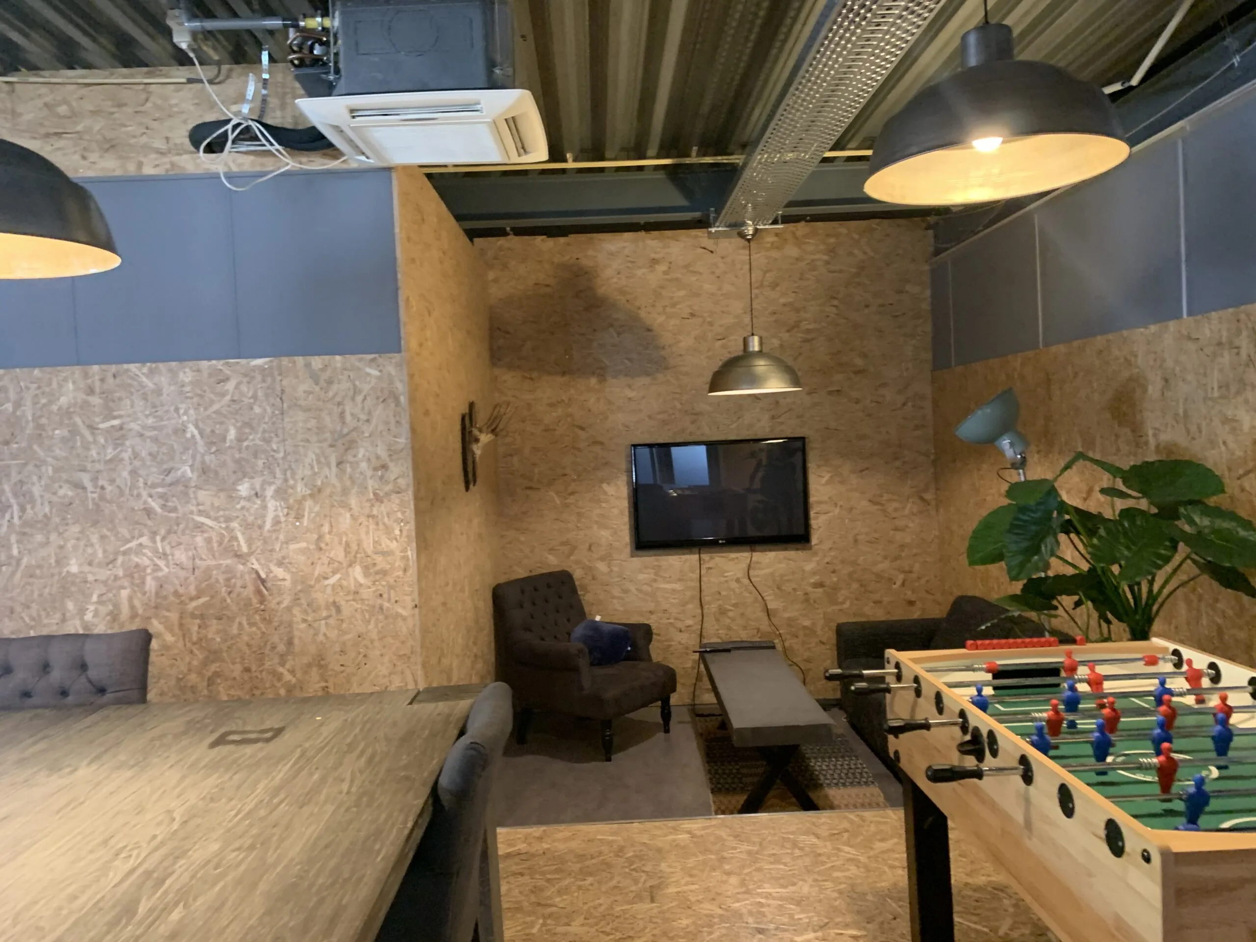 Ontspanningsruimte met tafelvoetbaltafel, zithoek, planten en wandtelevisie in een industriële setting.