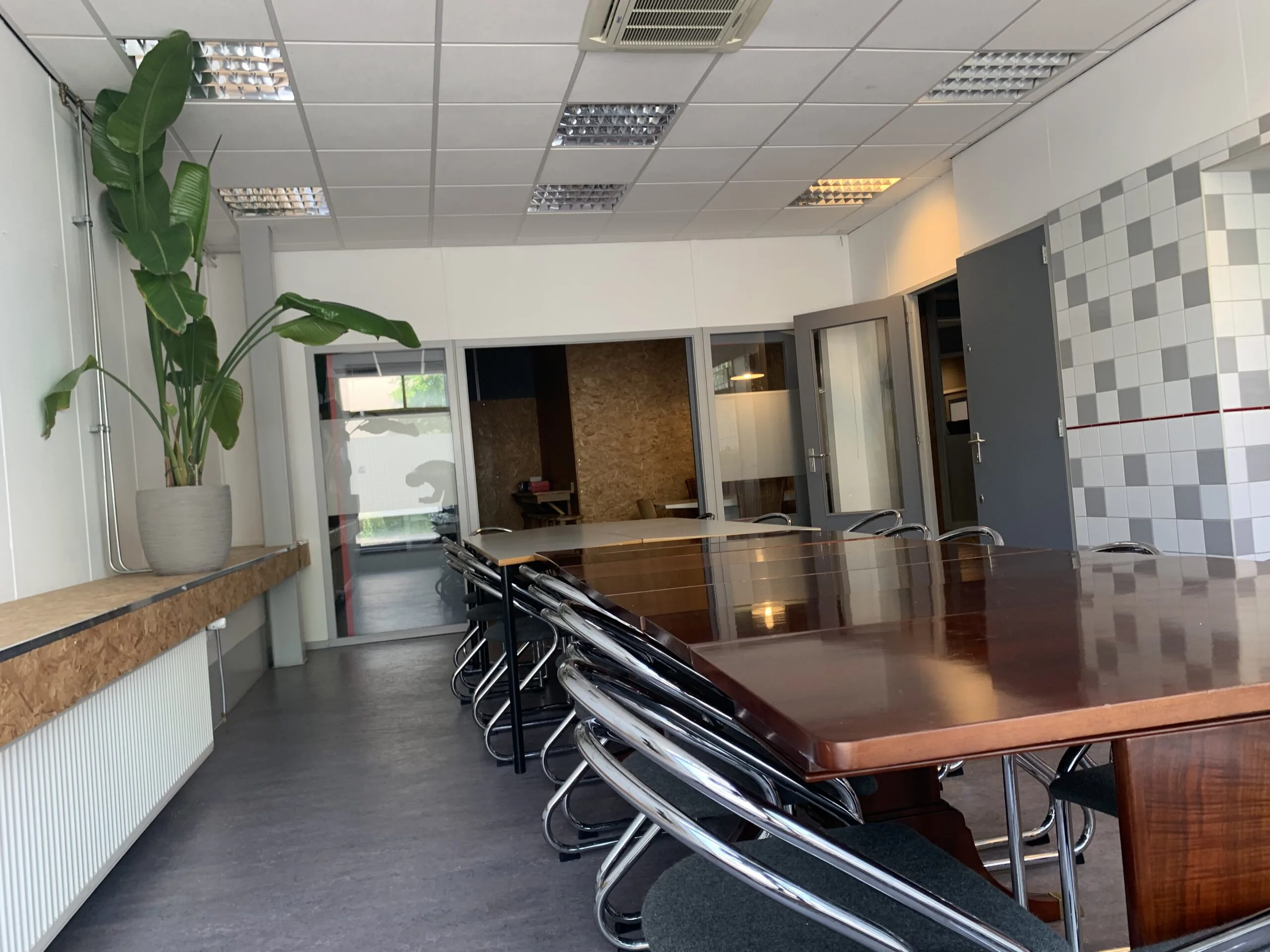 Vergaderruimte met een lange houten tafel, metalen stoelen, een grote plant en zicht op een aangrenzende kamer.
