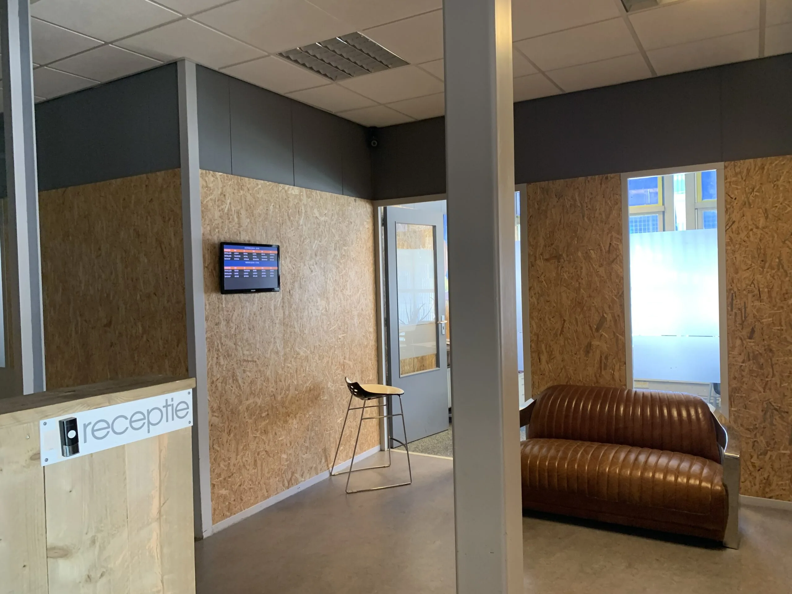Moderne ontvangsthal met houten receptiebalie, leren bank en muur met monitor.