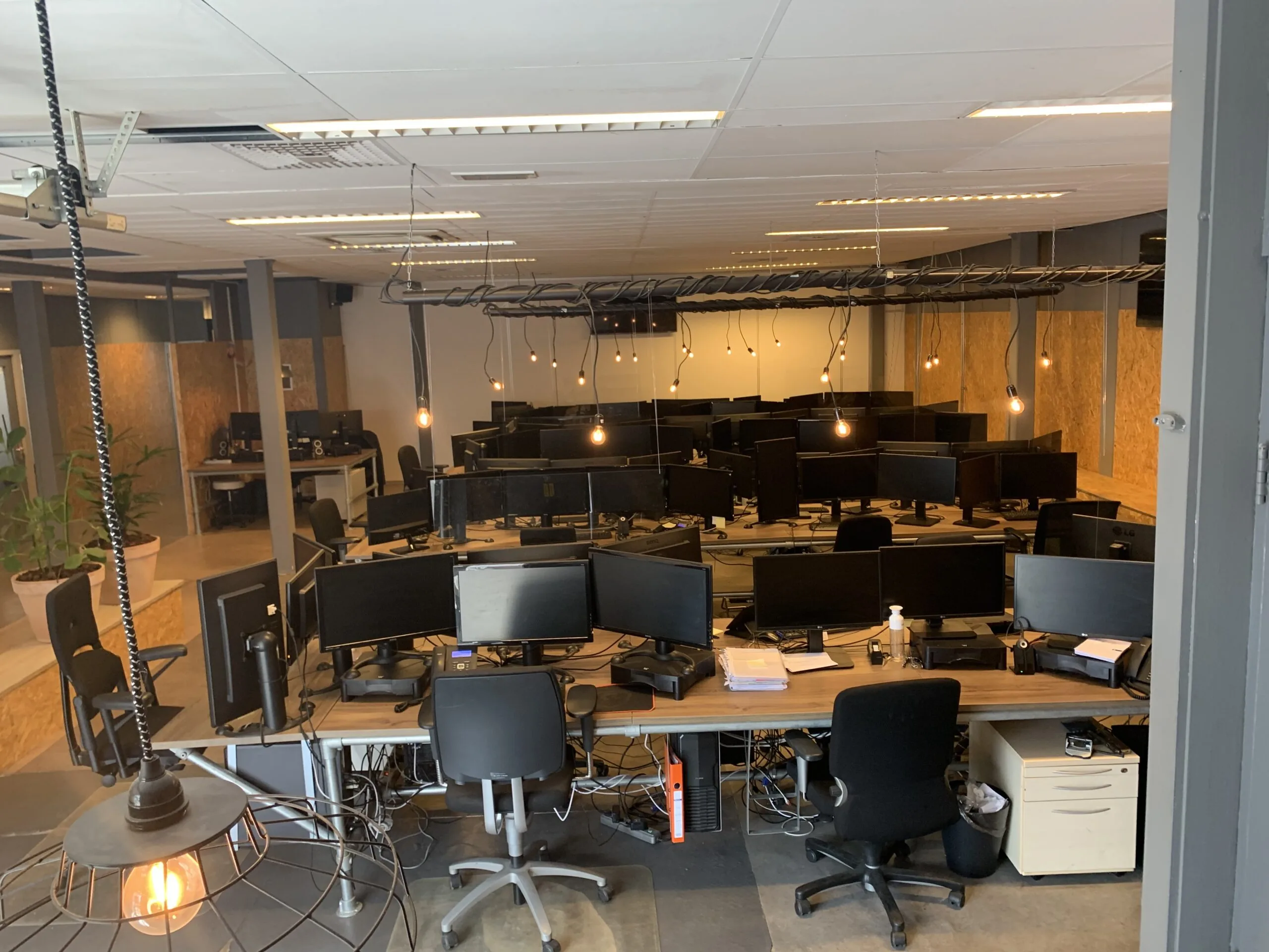 Open kantoorpand met meerdere werkplekken, bureaus vol computerschermen en industriële hanglampen.
