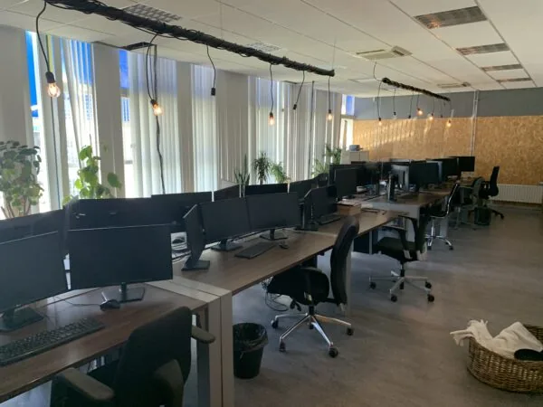 Modern kantoor met meerdere lege werkplekken, computerschermen en hanglampen langs grote ramen.