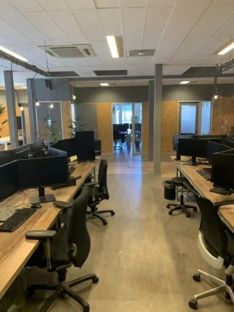 Moderne open kantoorruimte met bureaus, computerschermen en bureaustoelen, gezien vanaf het gangpad richting een vergaderruimte met glazen deuren.