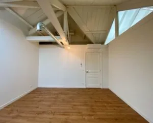 Lichte, lege kamer met houten vloer, wit geschilderde balken en een zichtbare ventilatiebuis aan het plafond.