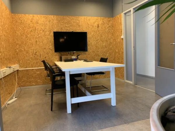 Vergaderruimte met een witte tafel, vier stoelen en een tv-scherm aan een wand van OSB-hout.