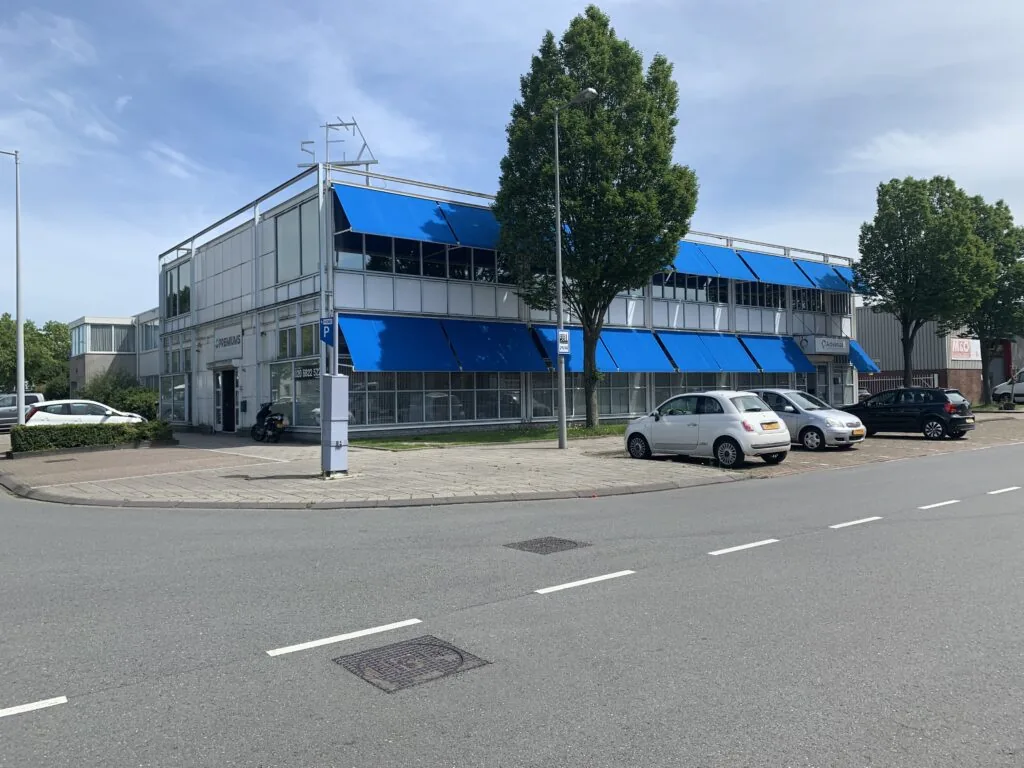 Kantoorgebouw met blauwe zonneschermen en geparkeerde auto's aan een rustige straat.