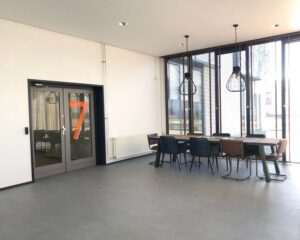 Moderne vergaderruimte met lange tafel, gemixte stoelen en grote ramen, naast een deur met het cijfer 7.