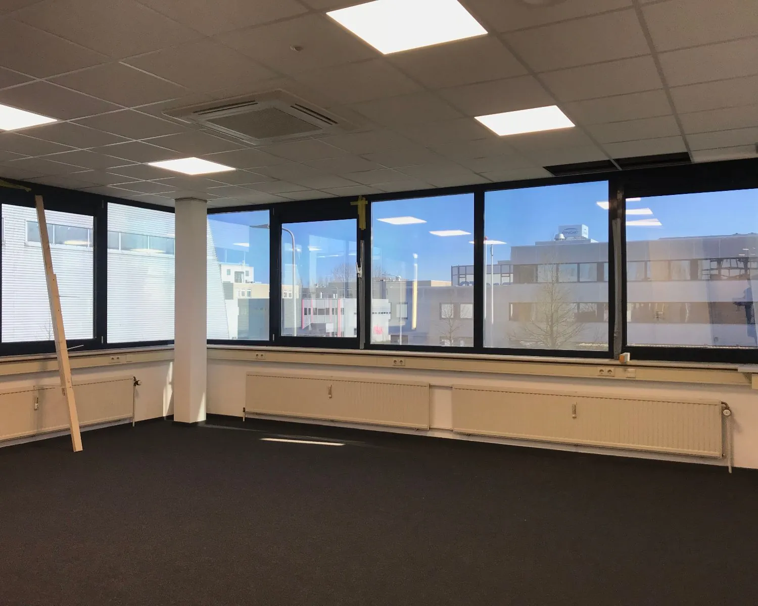 Leeg kantoor met systeemplafond, grote ramen en uitzicht op een modern bedrijfsgebouw.