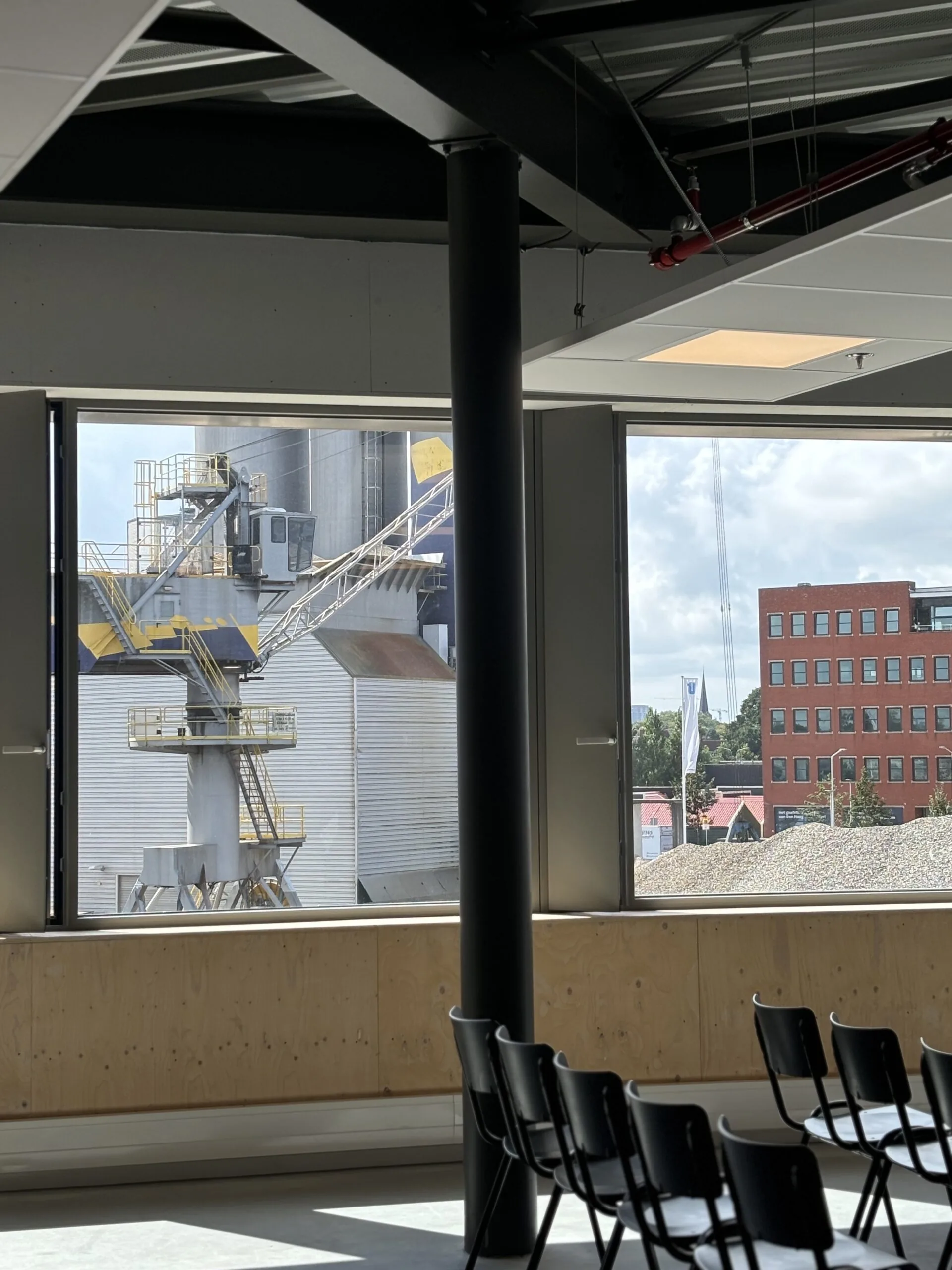 Zicht vanuit een modern ingerichte binnenruimte met stoelen op een industriële installatie en een rood kantoorgebouw buiten.