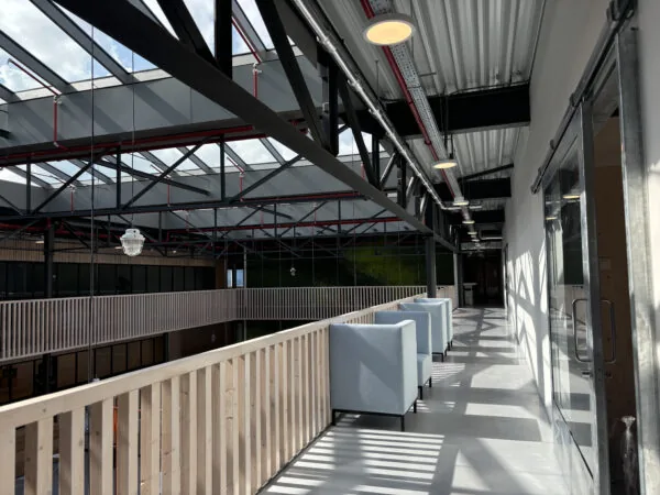 Zonovergoten gang met glazen dak, houten balustrade en lichtblauwe zitbanken in een modern industrieel gebouw.