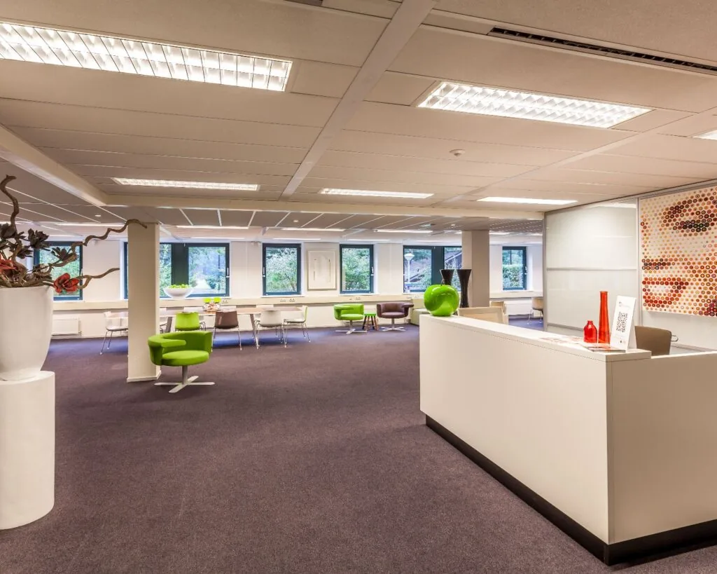Moderne kantoorruimte met een receptiebalie, kleurrijke zitplaatsen en grote ramen die daglicht binnenlaten.