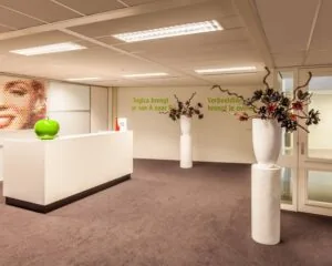 Moderne kantoorentree met een witte balie, kunstbloemstukken in hoge vazen en een inspirerende quote op de muur.