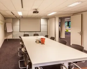 Vergaderruimte met een lange tafel, stoelen, een flip-over, een beamer en een open deur naar een aangrenzende ruimte.