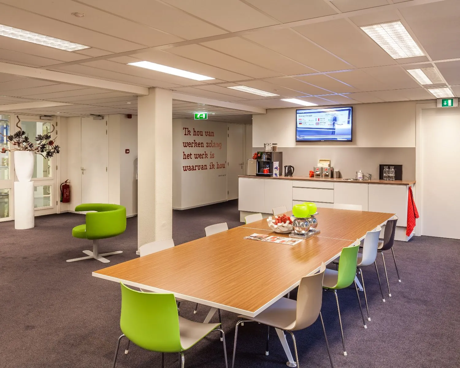 Moderne kantoor-kantine met een lange tafel, kleurrijke stoelen, koffiecorner en een televisiescherm aan de muur.
