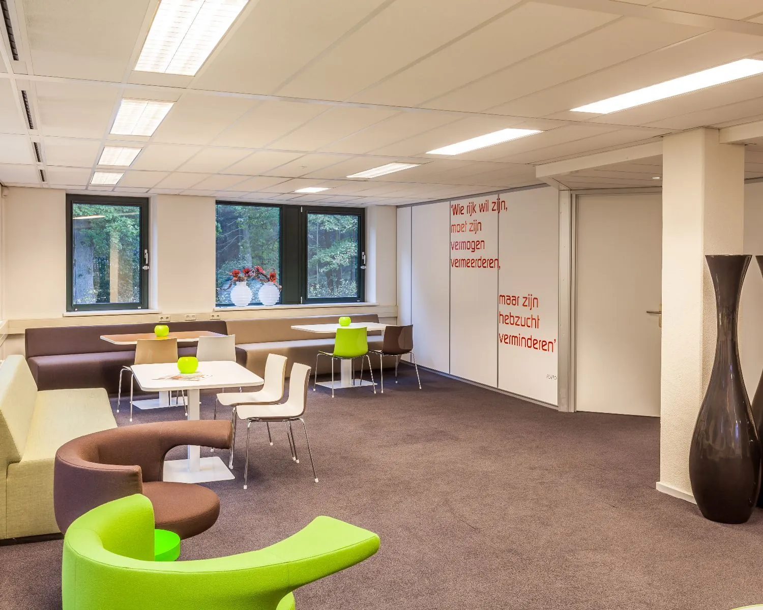 Moderne, licht ingerichte kantoorlounge met kleurrijke stoelen, tafels en grote ramen met uitzicht op bomen.