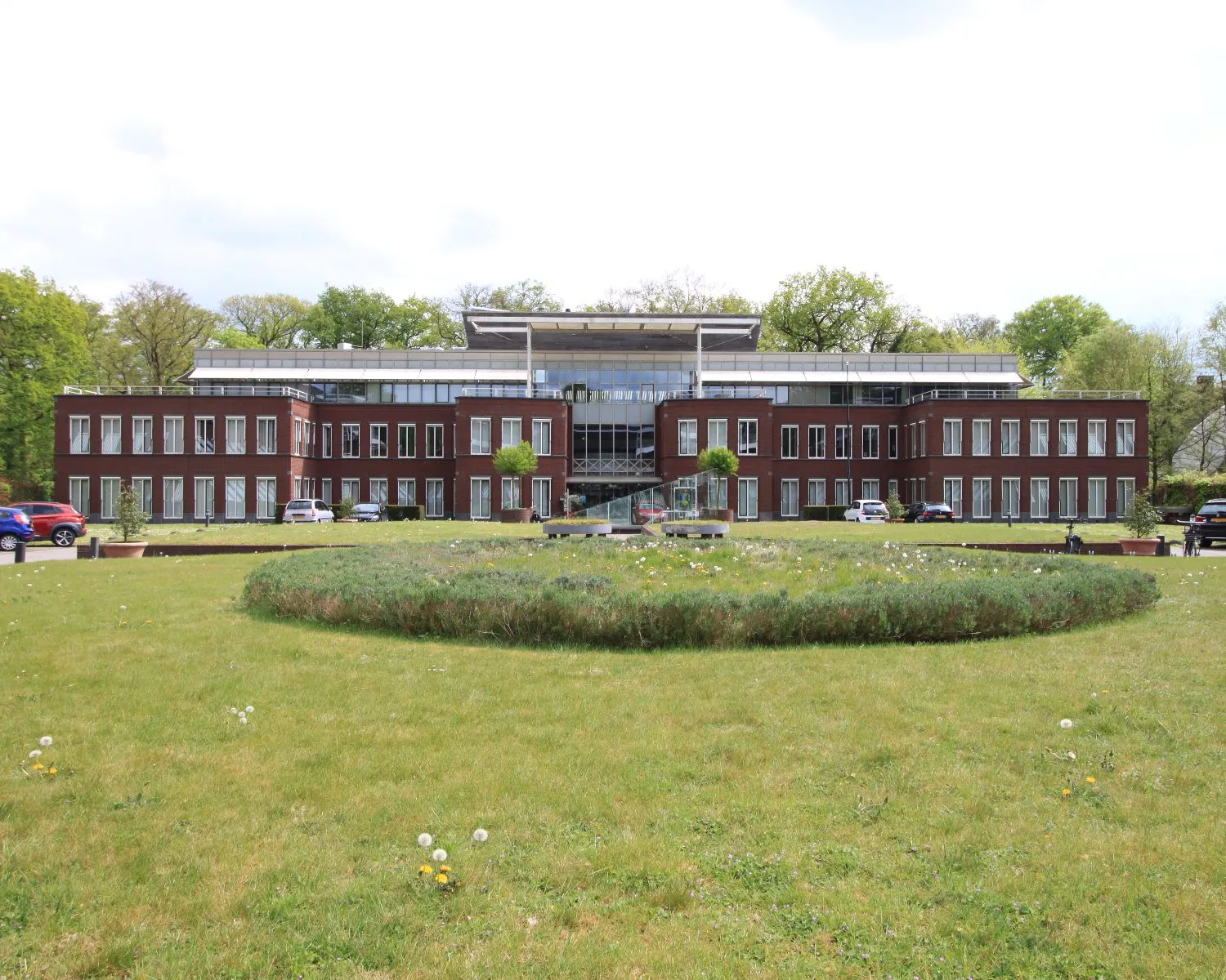 Modern kantoorgebouw met grote ramen en een ronde bloementuin op de voorgrond.