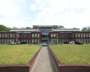 Modern kantoorgebouw met rode bakstenen gevel en glazen entree, gezien vanaf een stenen looppad door een grasveld.