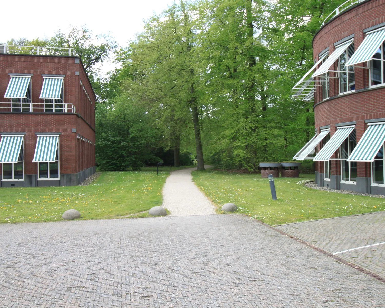 Pad tussen twee rode bakstenen kantoorgebouwen met groen-wit gestreepte zonweringen en groene bomen op de achtergrond.