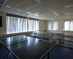 Kantineruimte met een tafeltennistafel, houten picknickbanken en veel daglicht door grote ramen met verticale lamellen.