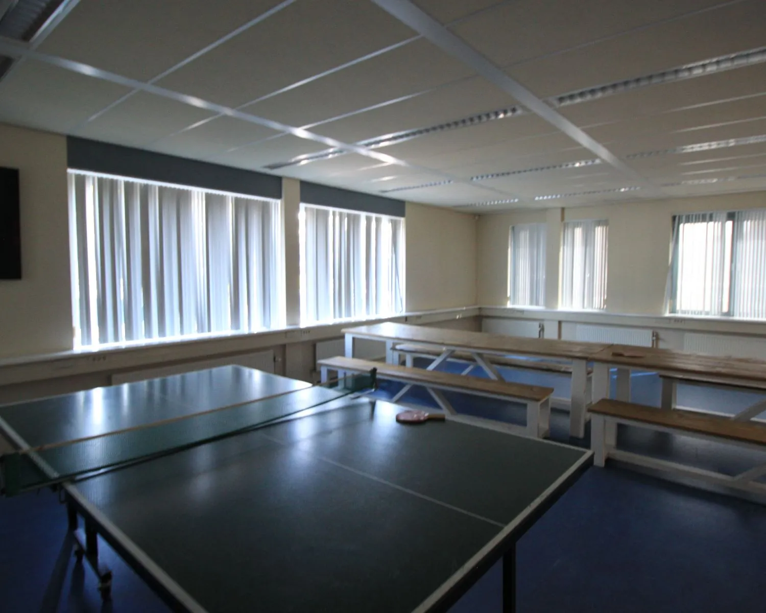 Kantineruimte met een tafeltennistafel, houten picknickbanken en veel daglicht door grote ramen met verticale lamellen.