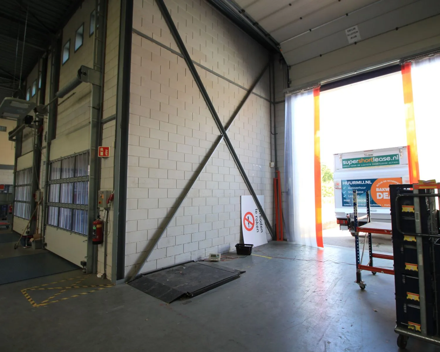 Oprijplaat en magazijnkar nabij open laadruimte met een vrachtwagen van supershortlease.nl op de achtergrond.