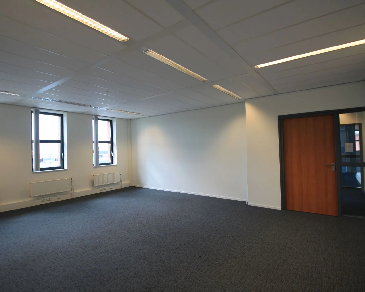 Leeg kantoor met drie ramen, grijze vloerbedekking en een gesloten houten deur.