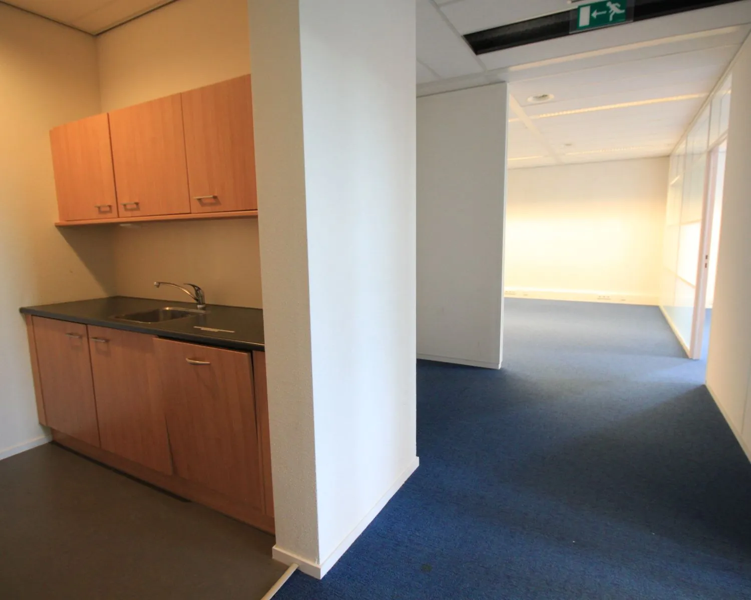 Kleine pantry met houten keukenkastjes in een kantoorruimte met blauw tapijt en witte wanden.