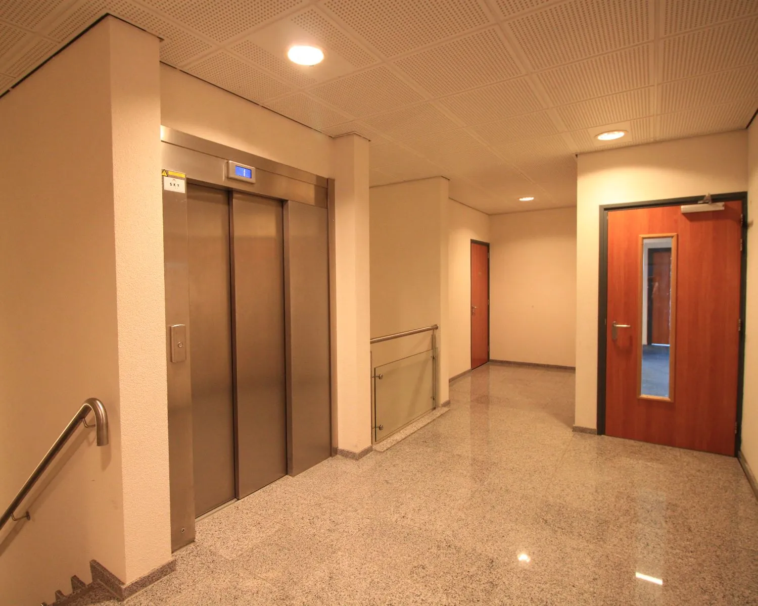 Verlichte binnenhal met een gesloten liftdeur, twee houten deuren en een trapleuning.