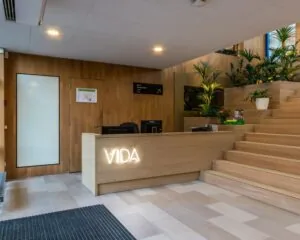 Receptiebalie met het opschrift "VIDA" in een moderne, houten lobby met trap en veel groene planten.