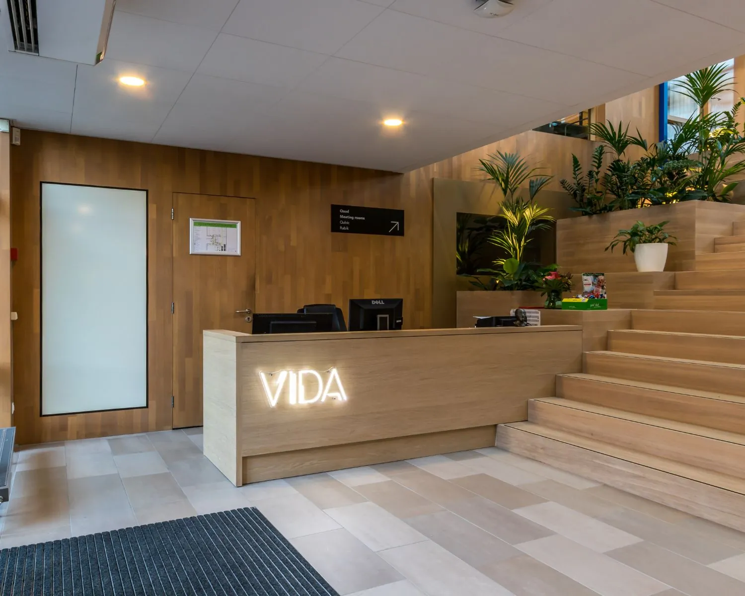 Receptiebalie met het opschrift "VIDA" in een moderne, houten lobby met trap en veel groene planten.