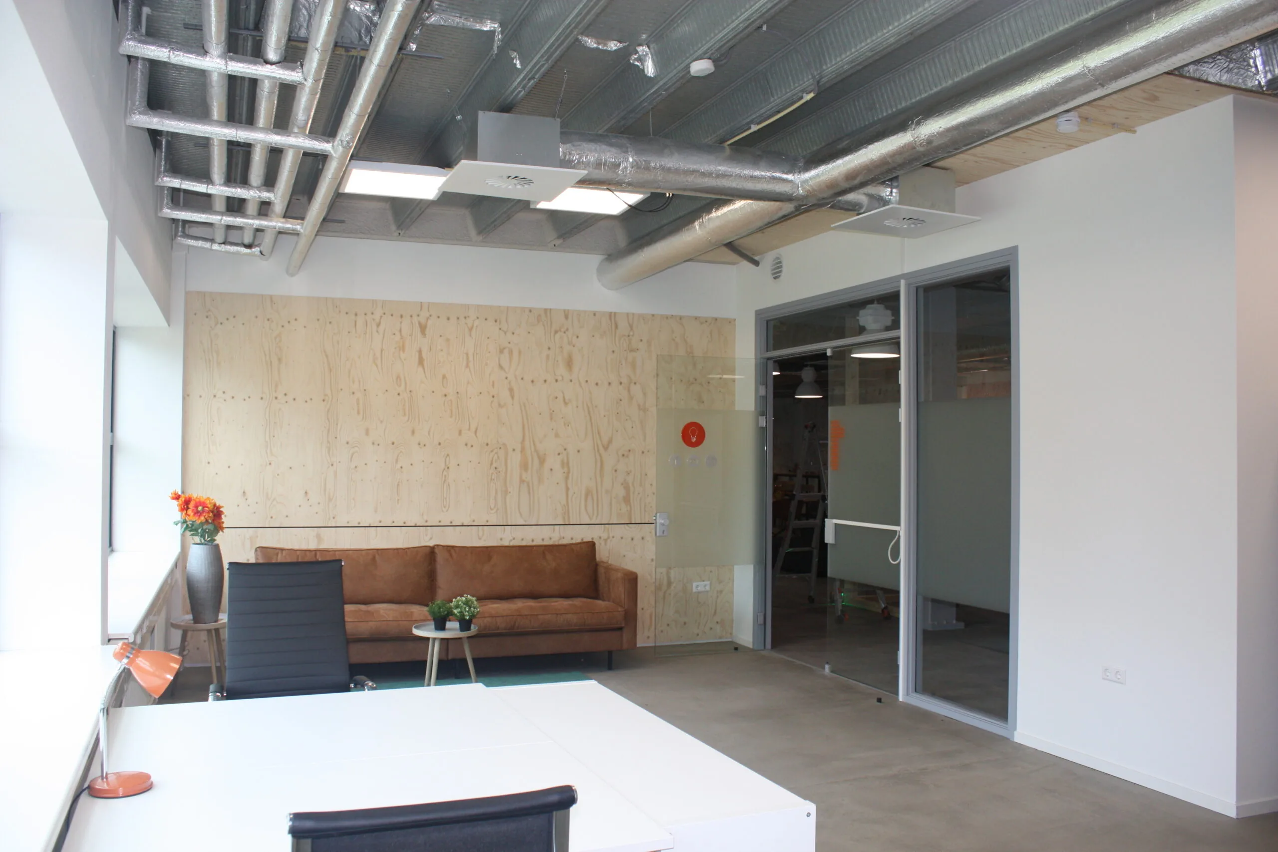 Modern kantoorinterieur met een leren bank, houten wandbekleding en zichtbaar plafond met ventilatiebuizen.