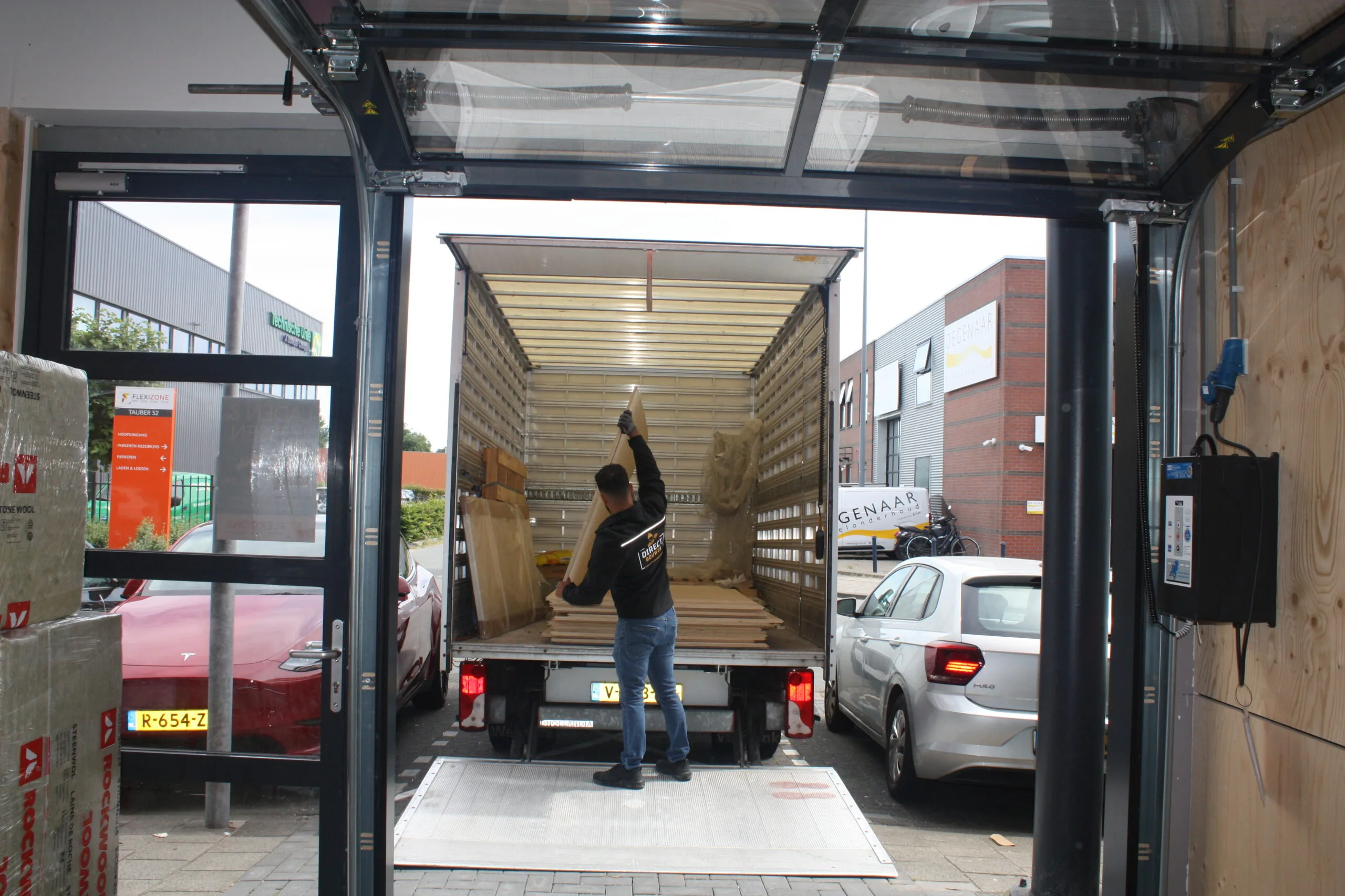 Man laadt houten platen in een vrachtwagen via een laadperron bij een bedrijfspand.
