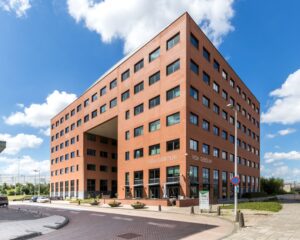 Roodbakstenen kantoorgebouw genaamd "Vida Gebouw" met moderne architectuur en een doorgang in het midden, onder een blauwe lucht met witte wolken.