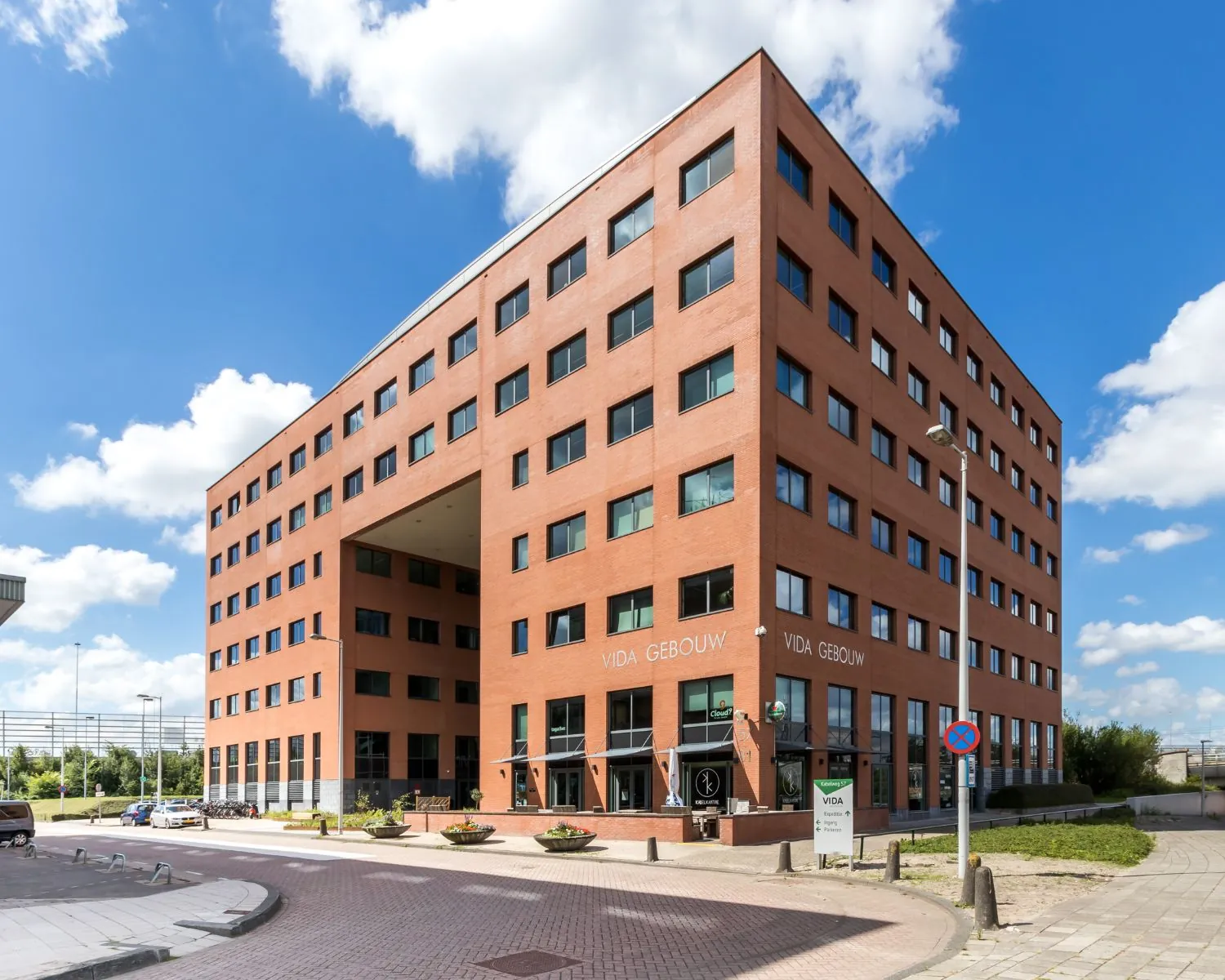 Roodbakstenen kantoorgebouw genaamd "Vida Gebouw" met moderne architectuur en een doorgang in het midden, onder een blauwe lucht met witte wolken.