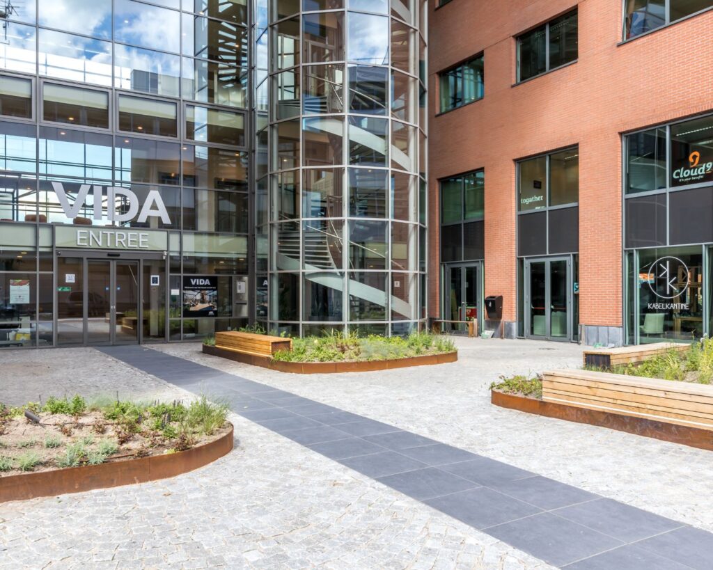 Entree van het VIDA-gebouw met glazen gevel, buitentrappenhuis en omliggend plein met banken en plantenbakken.