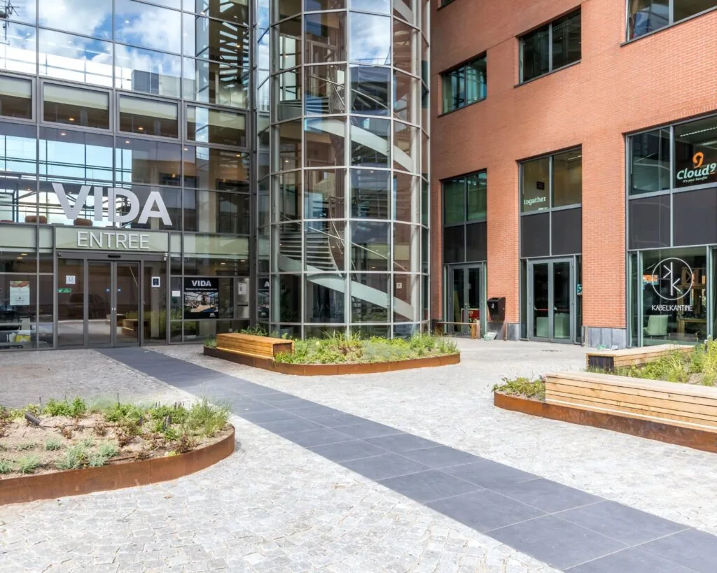 Entree van het VIDA-gebouw met glazen gevel, buitentrappenhuis en omliggend plein met banken en plantenbakken.
