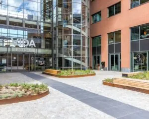 Entree van het VIDA-gebouw met glazen gevel, buitentrappenhuis en omliggend plein met banken en plantenbakken.